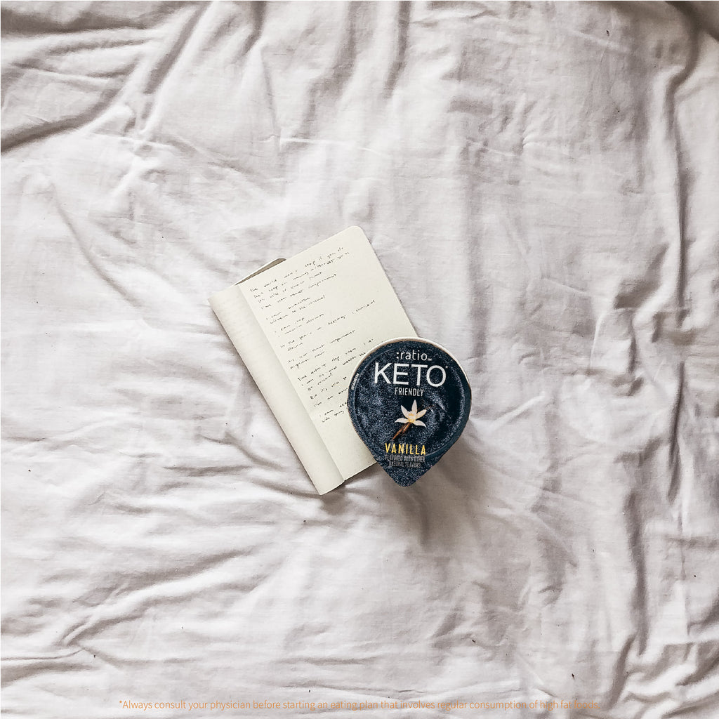 An open journal and a single serving cup of vanilla-flavored KETO Friendly Yogurt Cultured Dairy Snack on top of a bed.