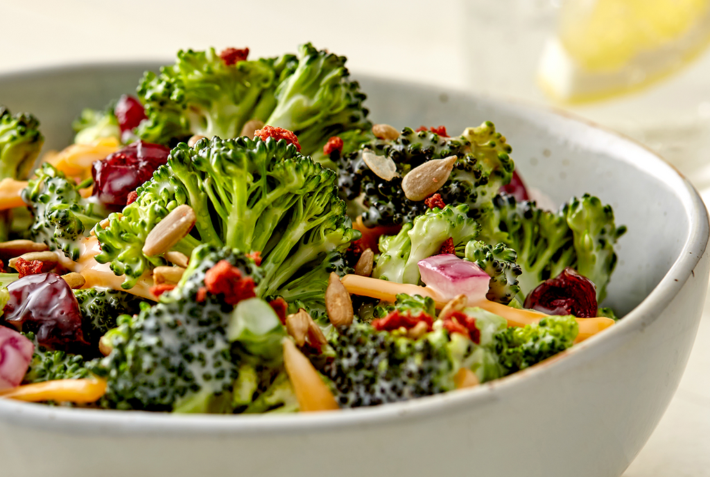 A bowl of broccoli salad containing red onions, nuts, dried fruit, and shredded cheese.