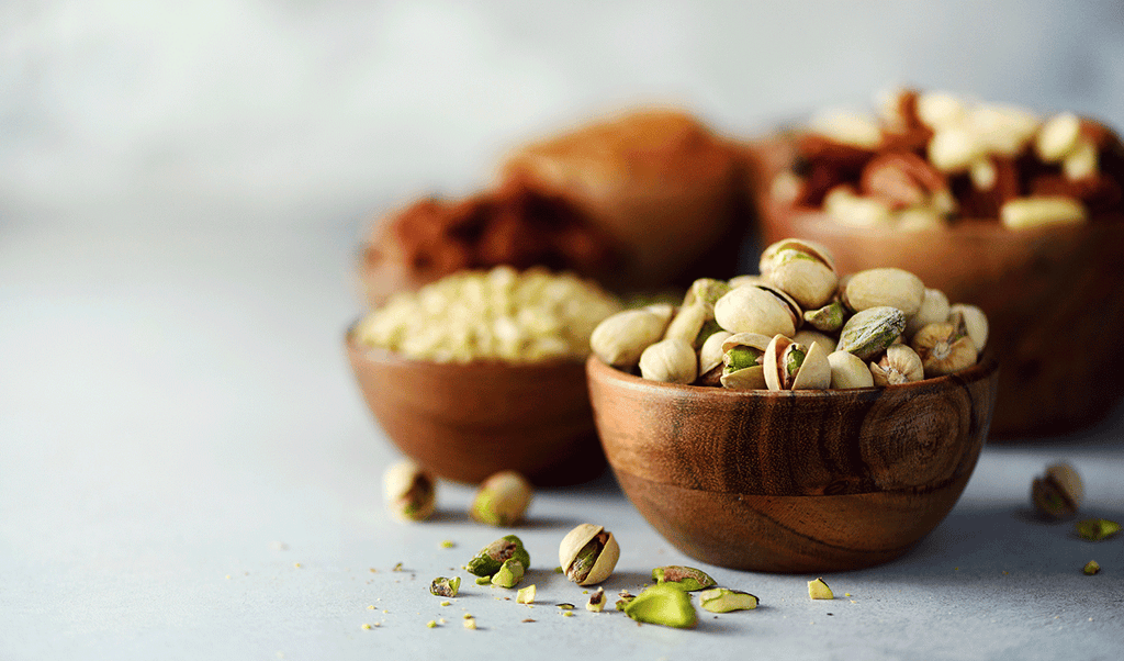 A small wooden bowl with pistachios on a light blue backdrop with assorted nuts in the background.