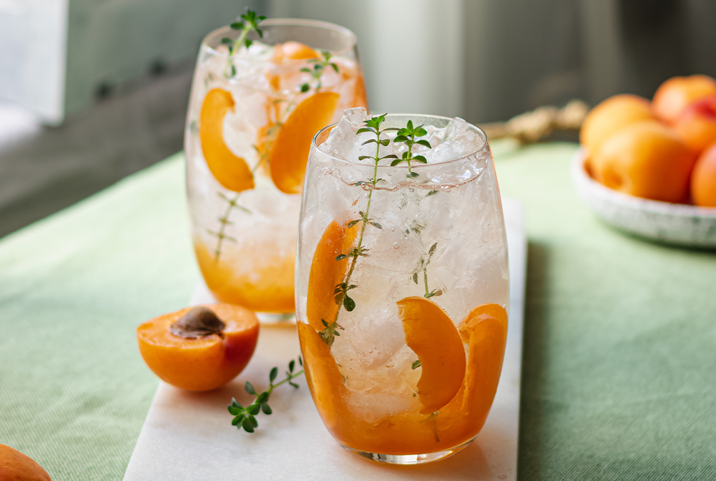 Two wine glasses filled with ice, a clear liquid, peach wedges, and garnishments. 