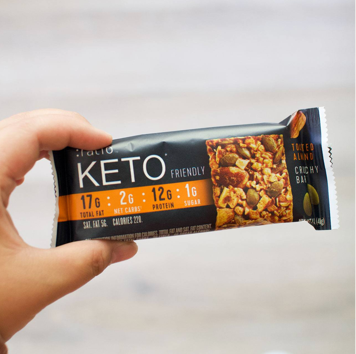 A hand holding a wrapped Ratio KETO Friendly Toasted Almond Crunchy Bar.