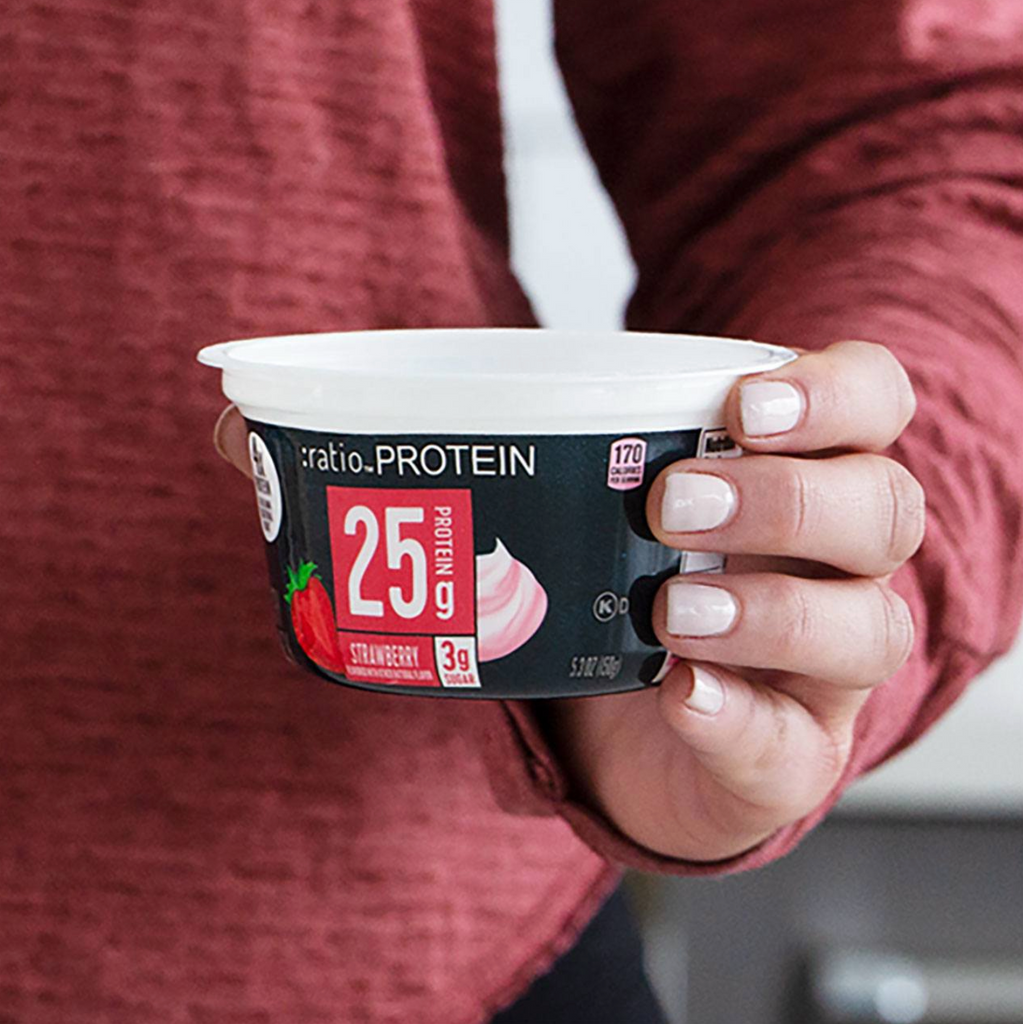 Person's hand displaying a cup of strawberry-flavored Ratio PROTEIN Yogurt Cultured Dairy Snack with 25g of Protein advertised on the label.